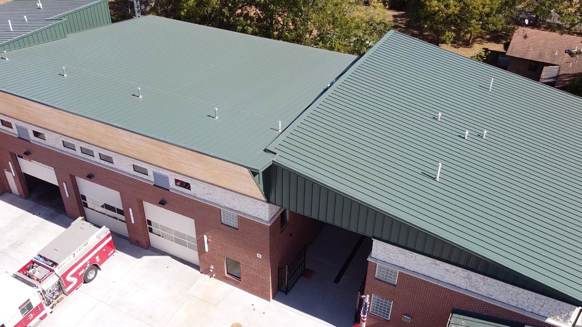 Aerial view of a brick fire station with a green metal roof. A fire truck is parked outside near several closed garage doors. Trees and another building are visible in the background.