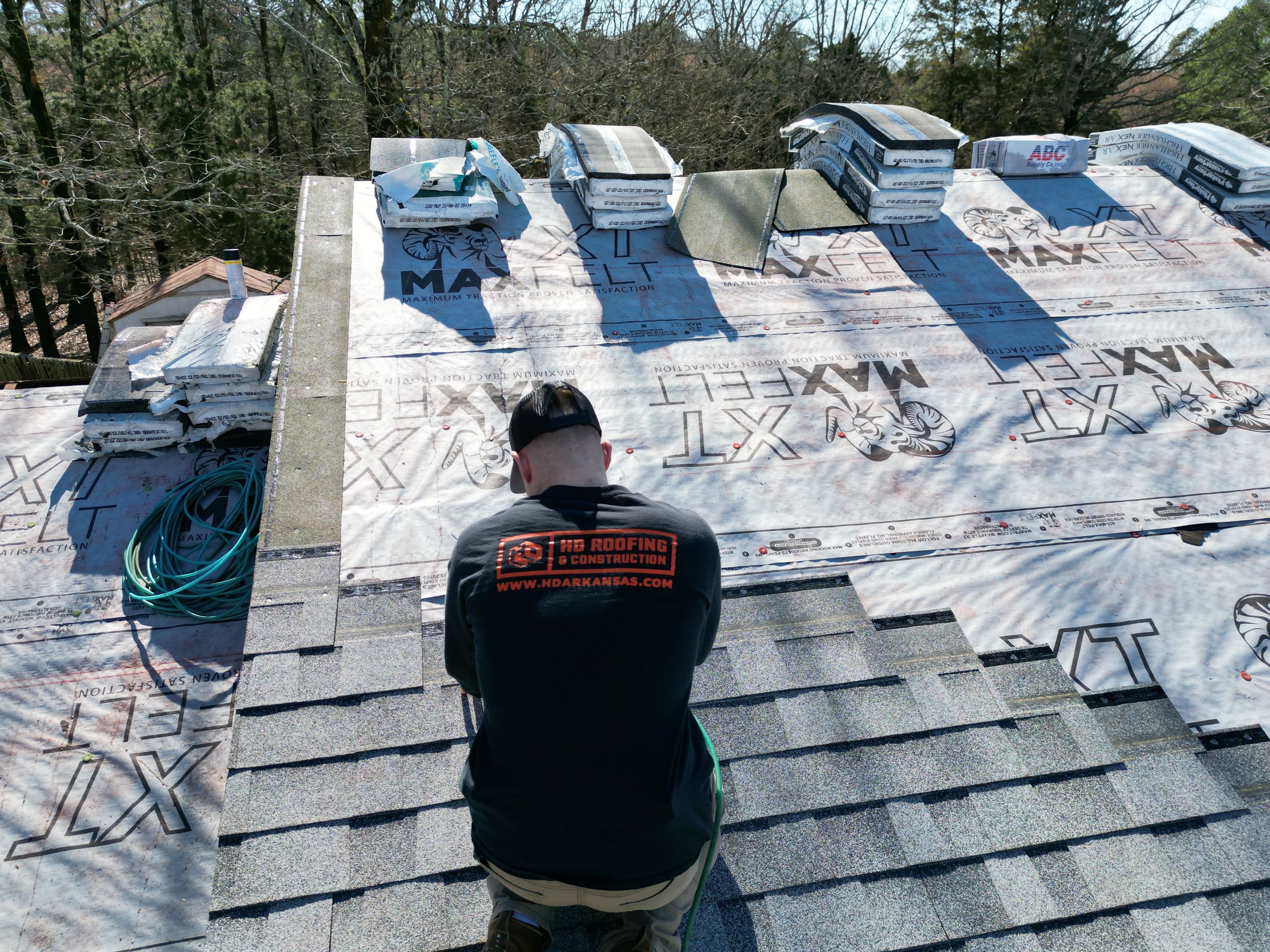 A roofing contractor in a black shirt installs shingles on a roof, surrounded by roofing materials and tools, with trees visible in the background on a sunny day.