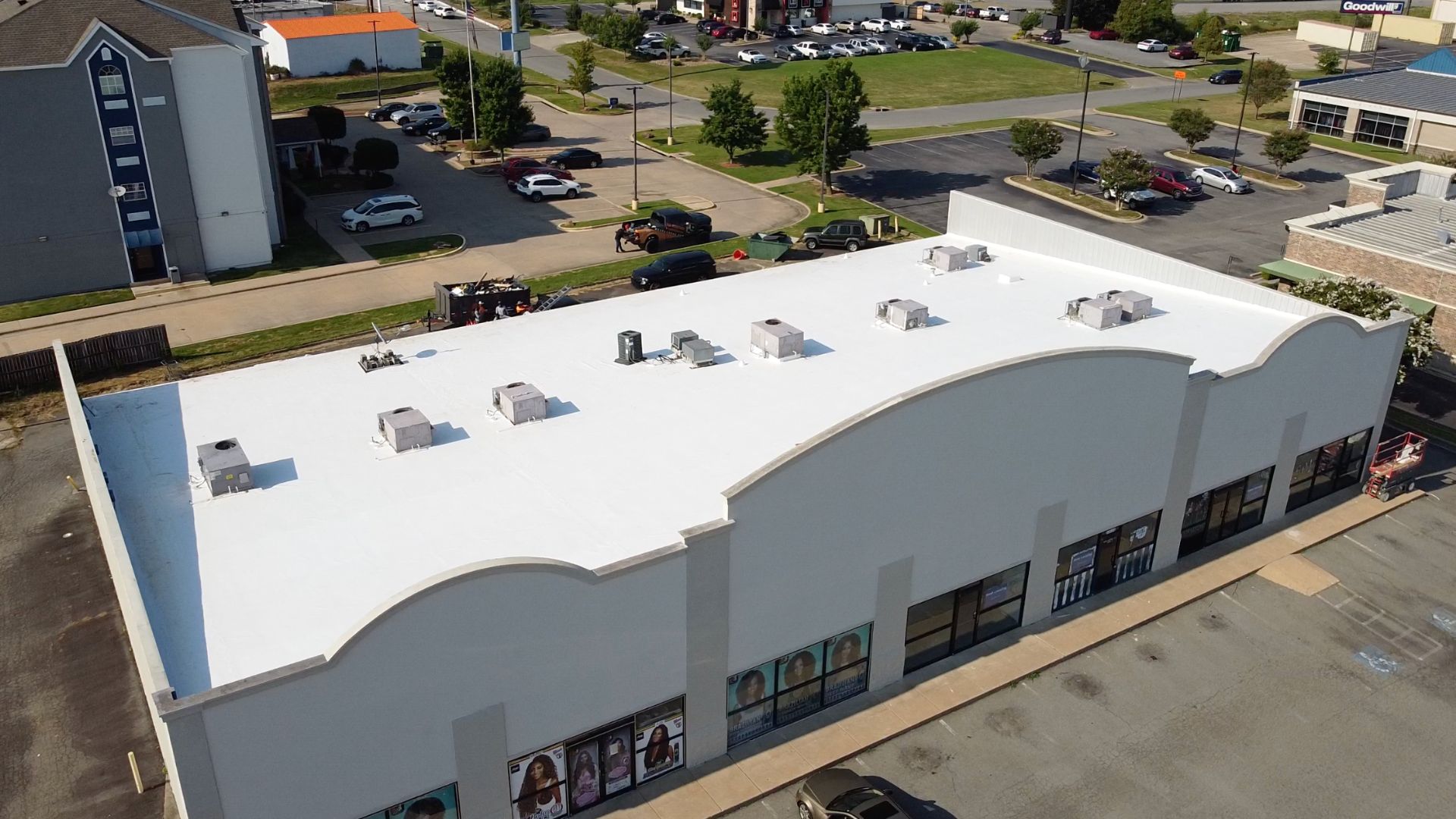 A large, single-story commercial building with a white, flat roof, surrounded by a parking lot with several cars and nearby businesses visible in the background.