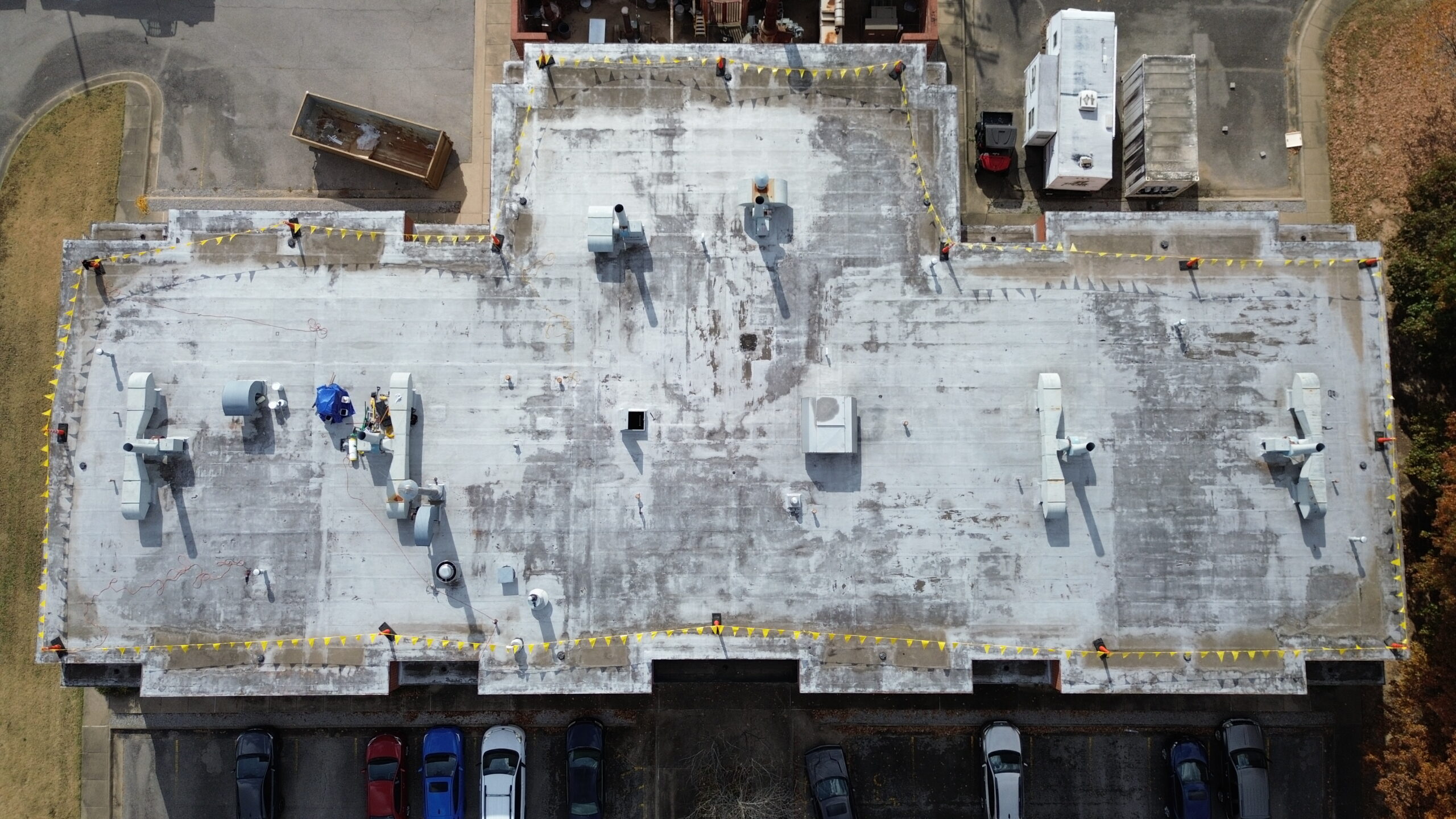 Aerial view of a large, rectangular, flat-roofed building with various HVAC units on top, surrounded by safety flags. Parked cars and some trees are visible around the building’s perimeter.