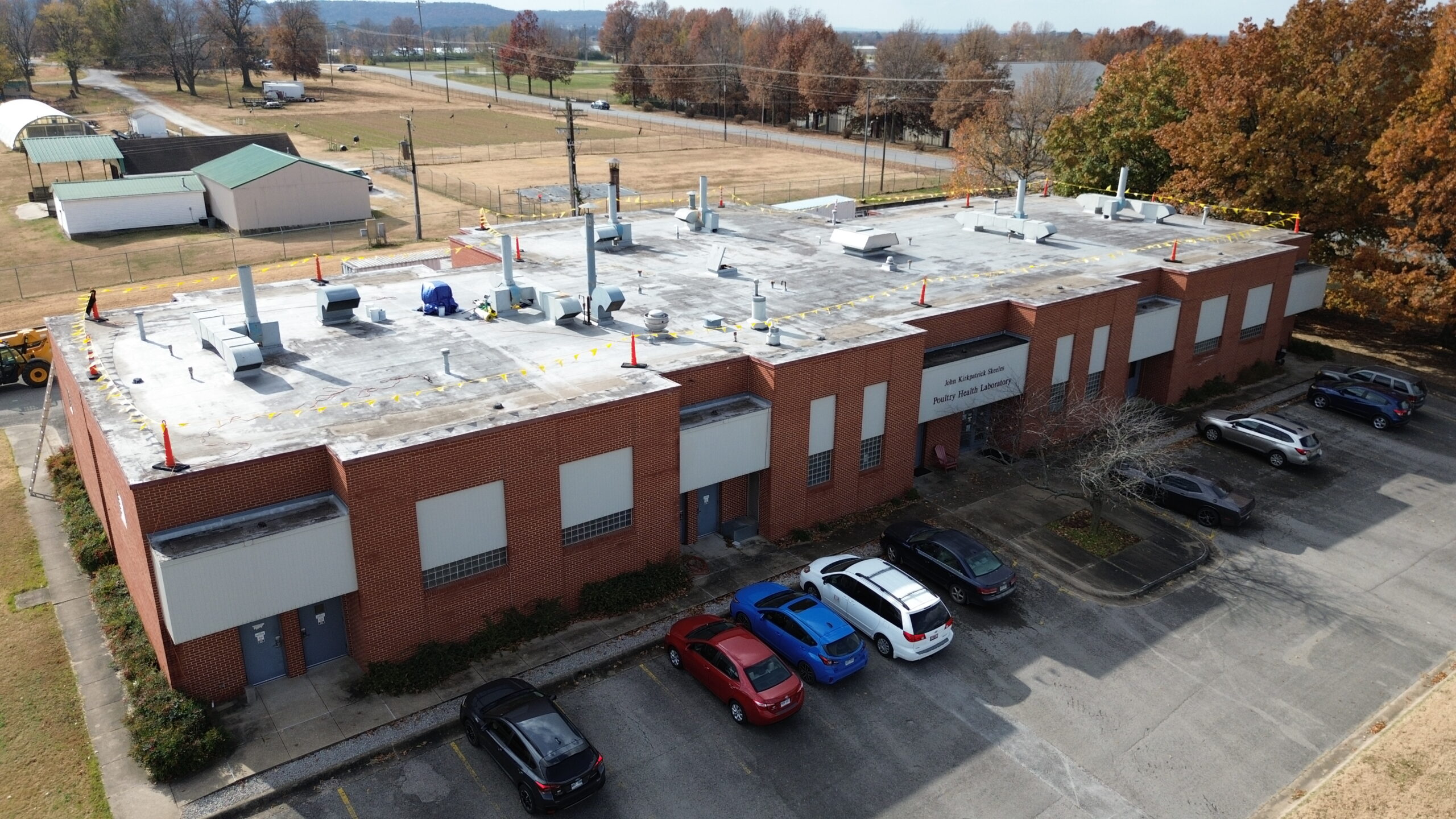 Aerial view of a flat-roofed brick building with several parked cars in front and autumn trees in the background. Rooftop equipment and safety cones are visible, with a mix of green and brown fields beyond.