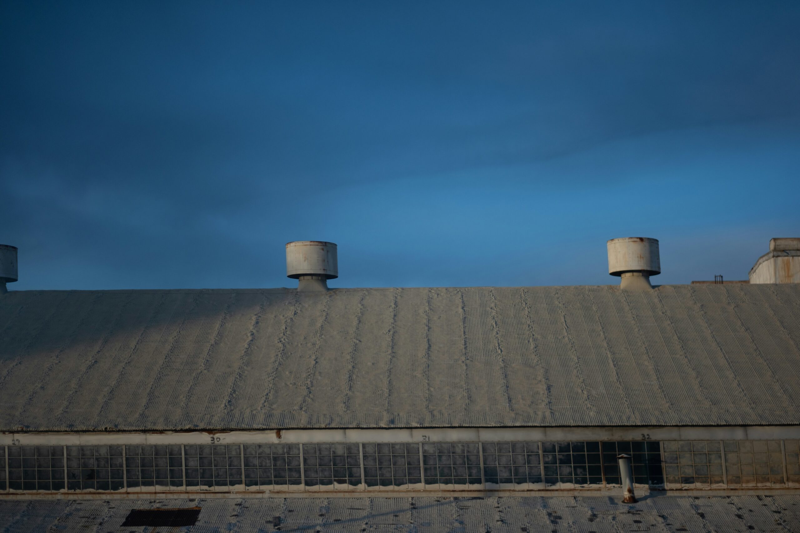 A large industrial building with a corrugated metal roof features two round roof ventilation units and rectangular glass windows below, all set beneath a clear blue sky.