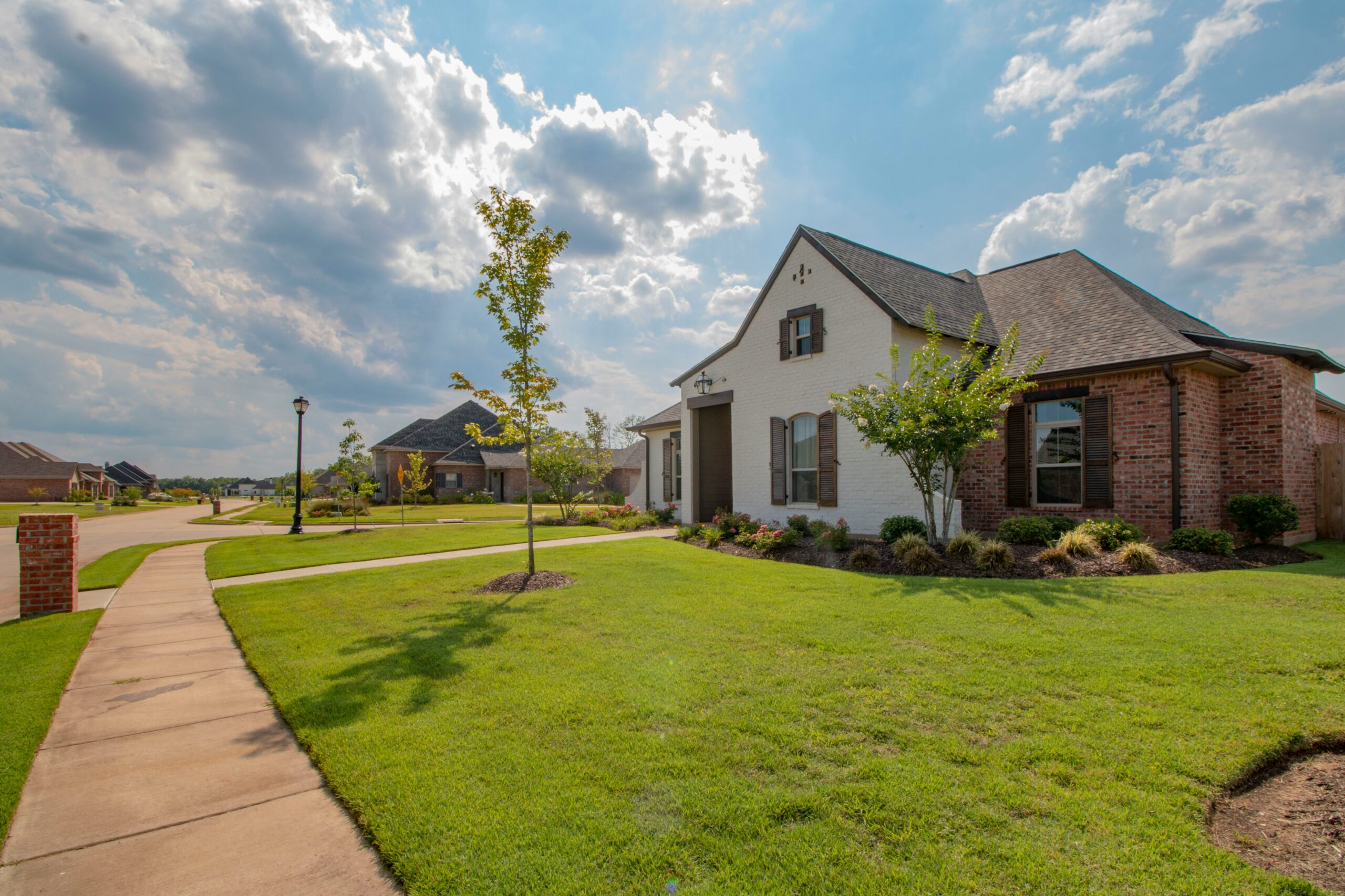 A suburban neighborhood with well-kept lawns, a sidewalk, and modern brick houses featuring energy-efficient roofing under a partly cloudy sky on a sunny day.