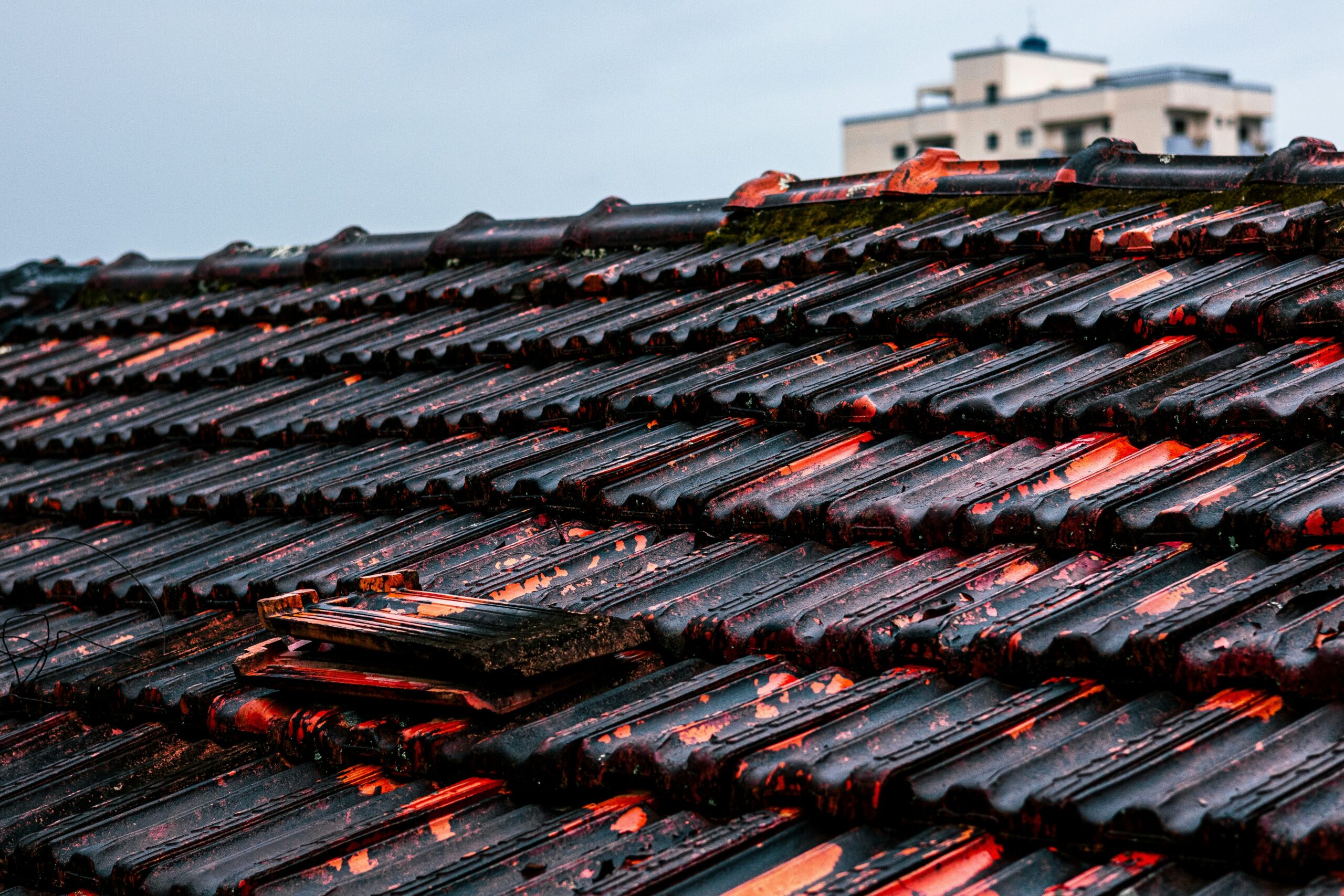 A close-up of a weathered tile roof with several broken and damaged tiles in need of roof repairs. In the background, a blurry apartment building is visible under a cloudy sky.
