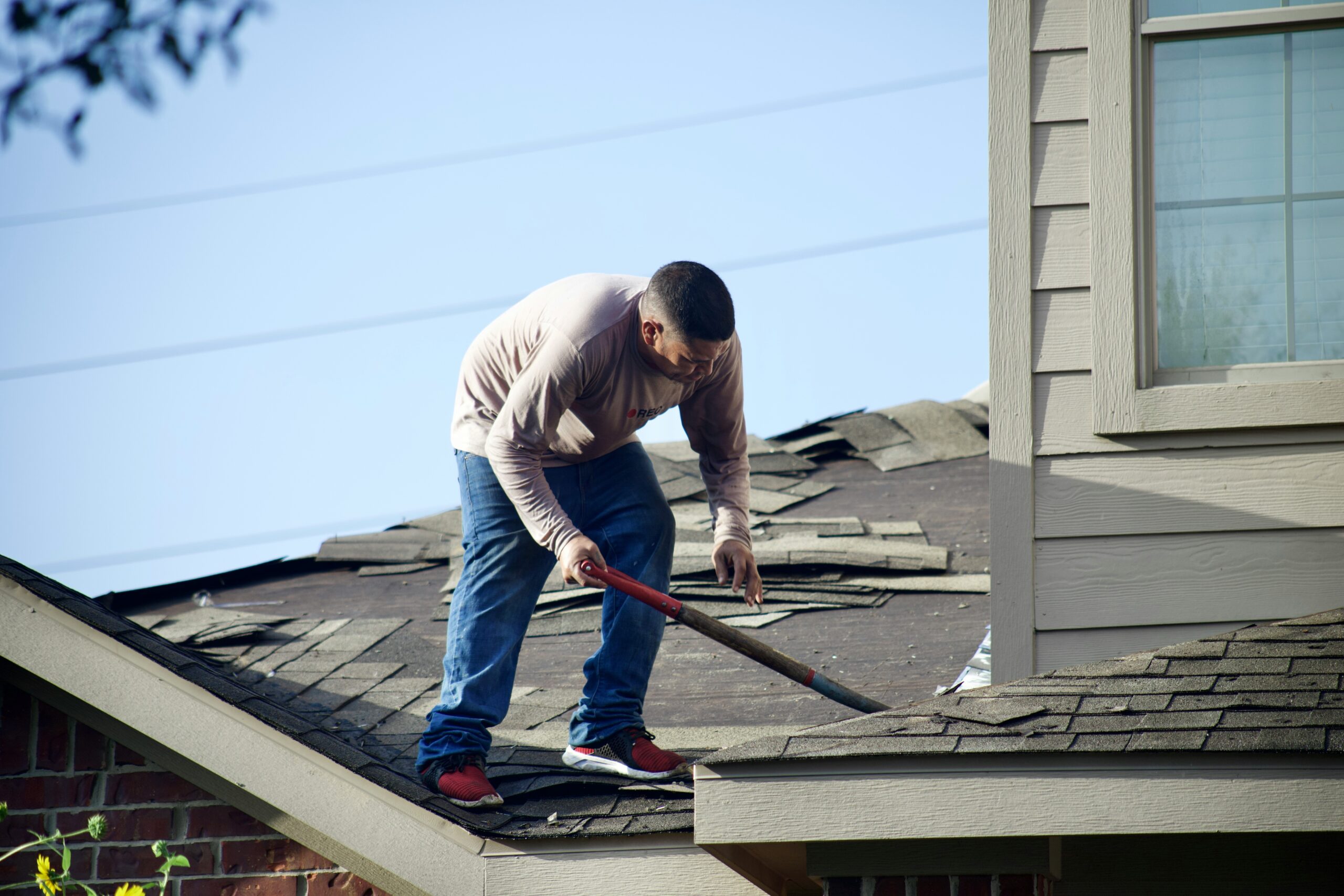 A person in jeans and a long-sleeve shirt uses a tool to perform roof leak repair, working on shingles under a clear blue sky.
