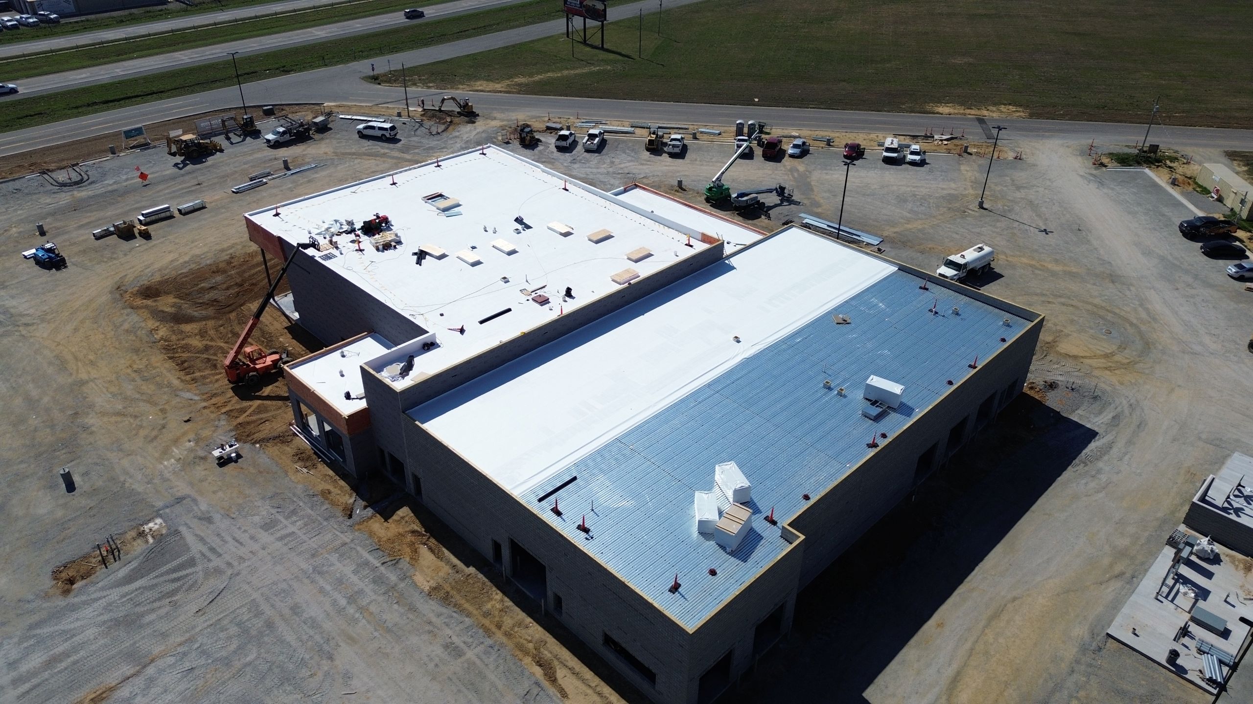 Aerial view of a large building under construction with an energy-efficient roofing system in white and blue, surrounded by dirt, construction equipment, and parked vehicles; a road and grassy field are visible in the background.