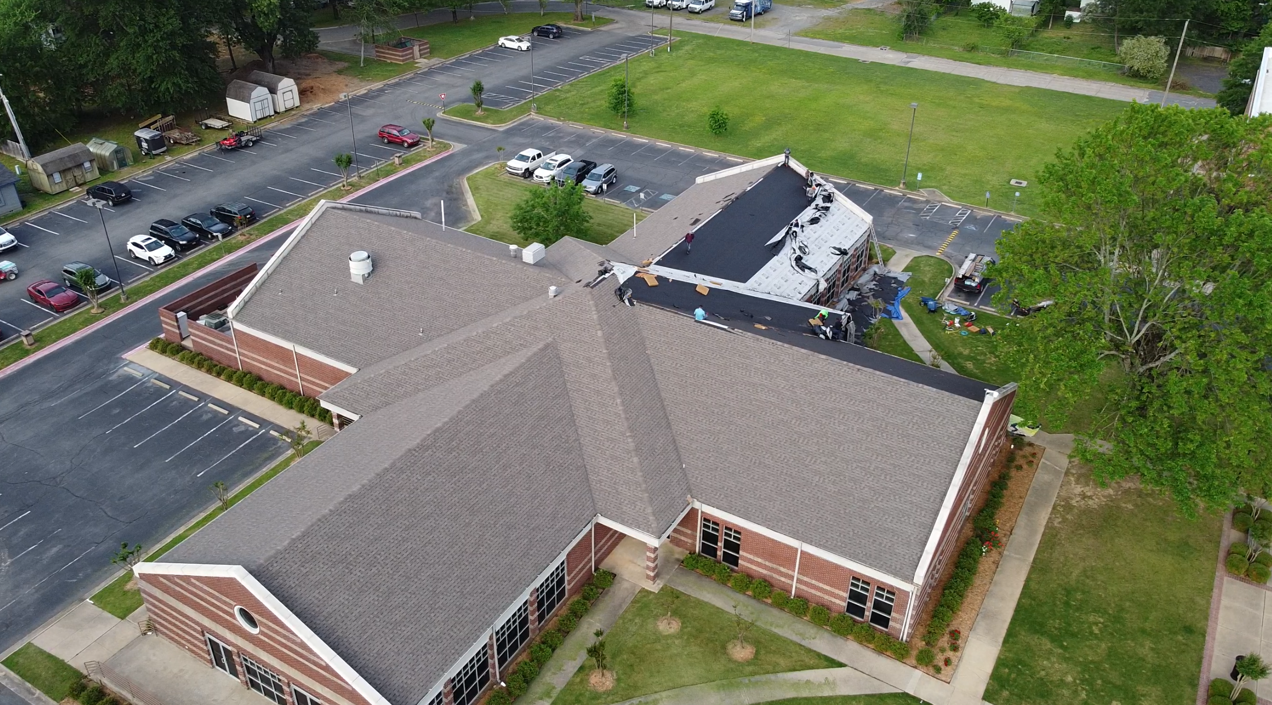 Aerial view of a large brick building with workers repairing common roof problems. Construction equipment is visible, and parking lots with several cars surround the building. Trees and grassy areas are nearby.
