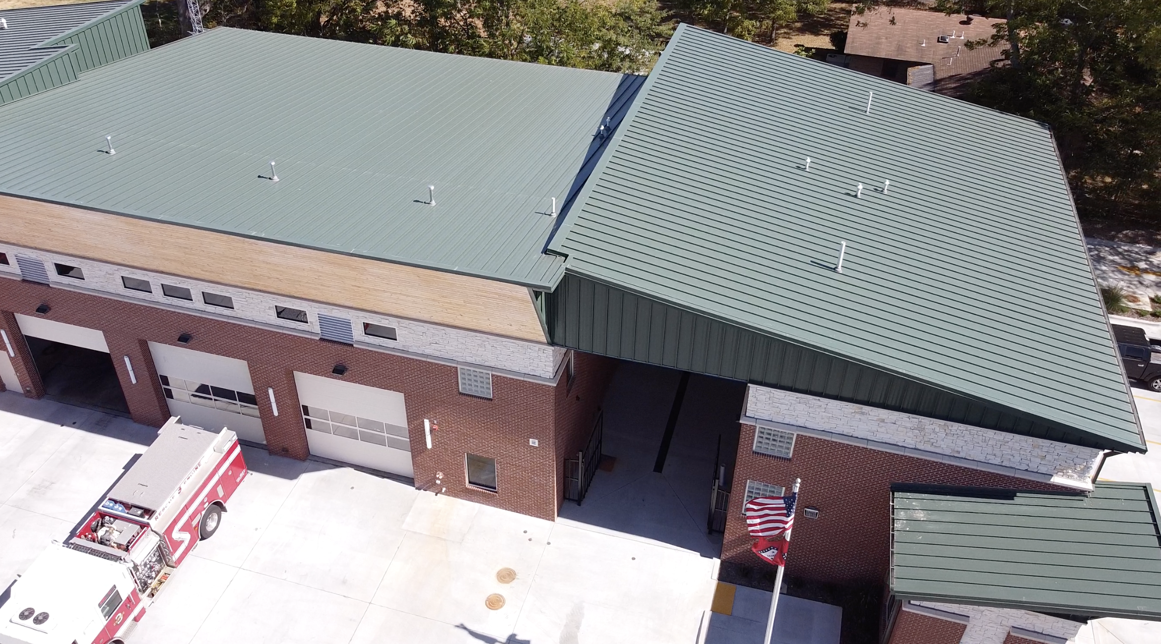 Aerial view of a modern brick fire station with green metal roofs, multiple garage doors, a fire truck parked outside, and an American flag waving near the entrance.