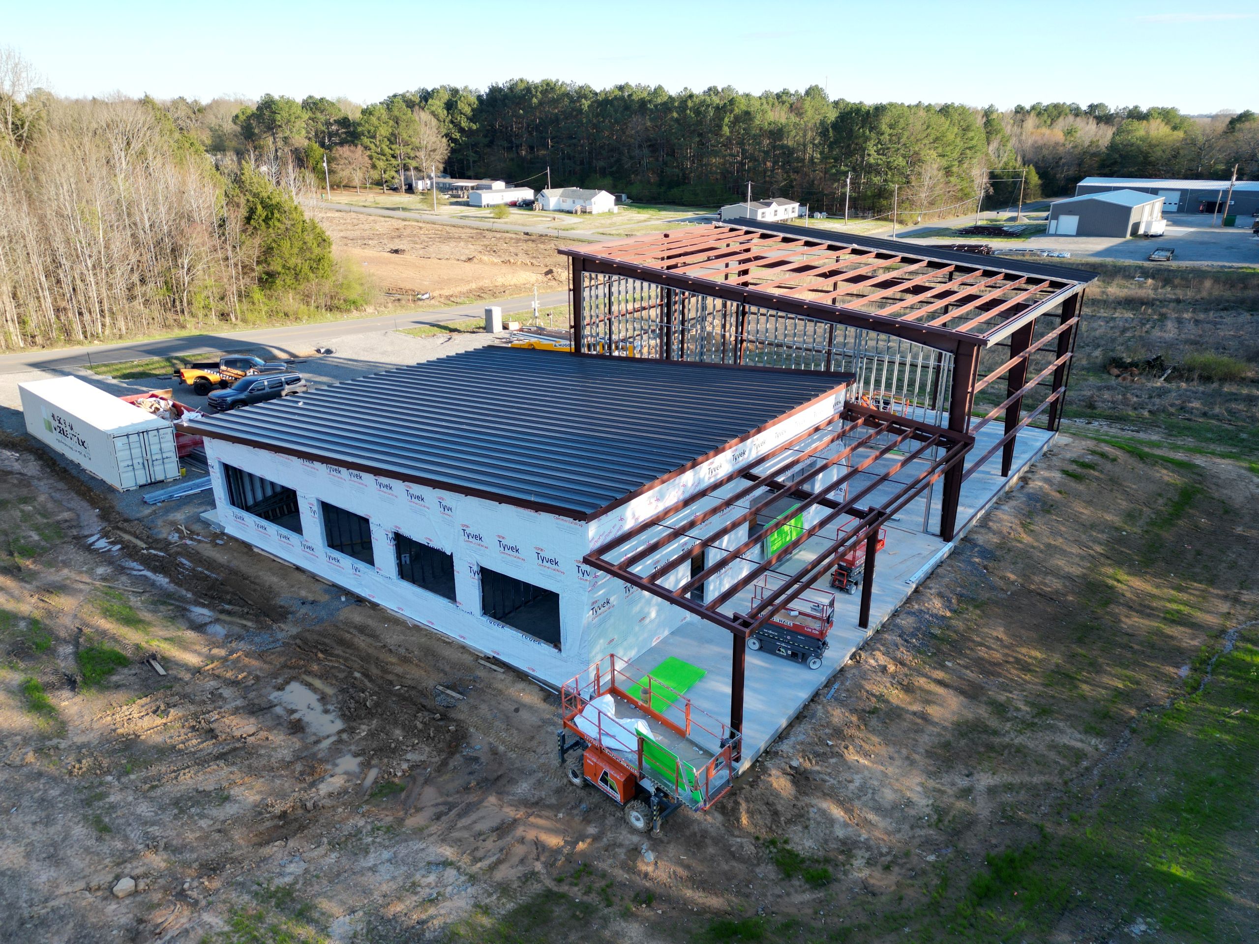 Aerial view of a commercial building undergoing roof repair services, featuring a partially completed steel frame and a section with a finished roof, surrounded by dirt and trees.