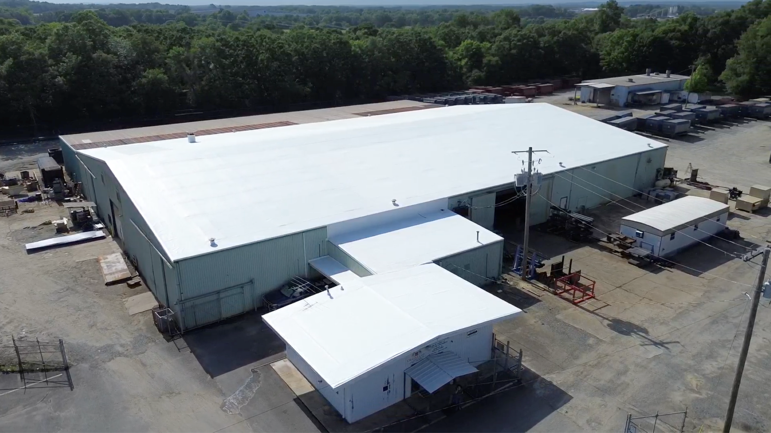 Preparing Manufacturing Facilities for Peak Storm Season A large industrial warehouse with a white roof is surrounded by trees and smaller buildings on a sunny day. Various equipment and vehicles are scattered around the paved lot.