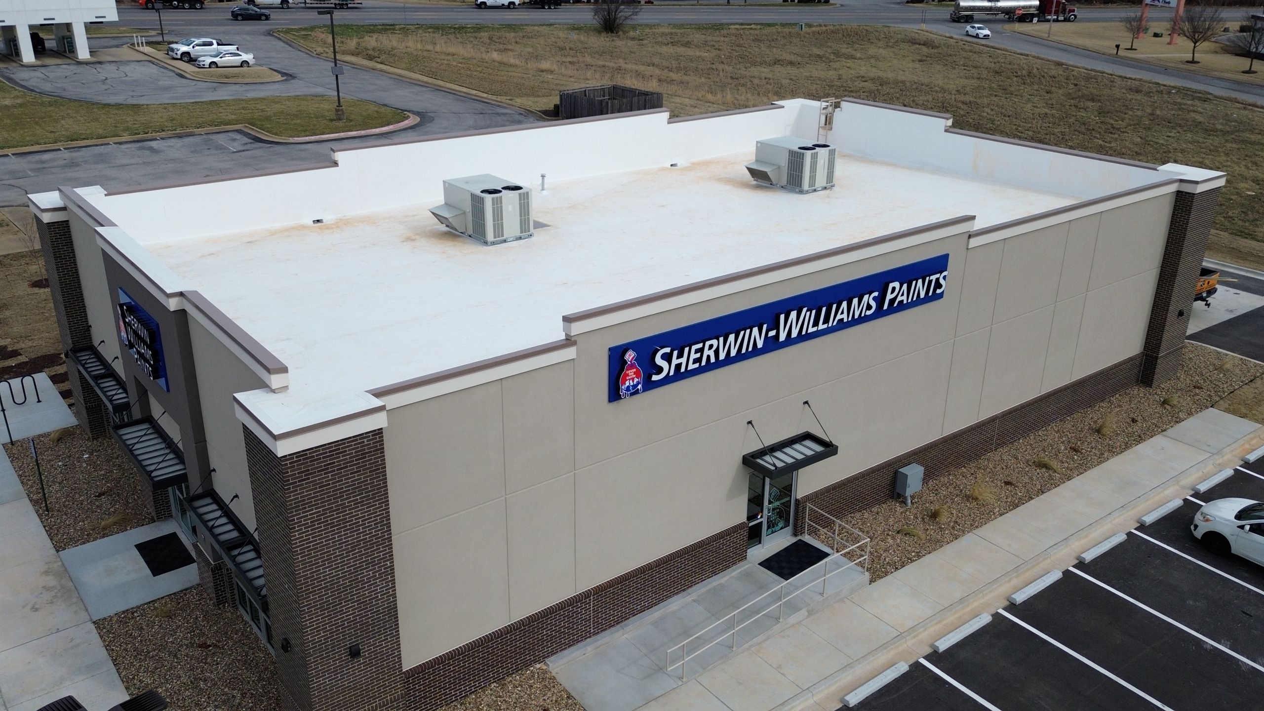 Why Experience Matters for Large Commercial Roof Replacements A commercial roof replacement of Sherwin-Williams Paints store with a flat white roof, beige walls, and a large blue sign. The parking lot is mostly empty, and nearby roads and grass surround the building.