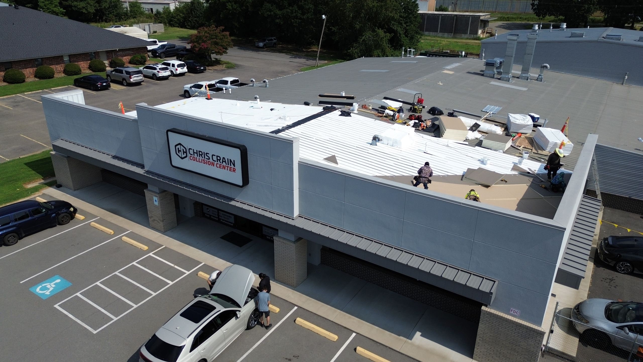 Aerial view of workers installing a new roof on the Chris Crain Collision Center building, with people attending to a car in the parking lot below, possibly showing commercial roof leaks